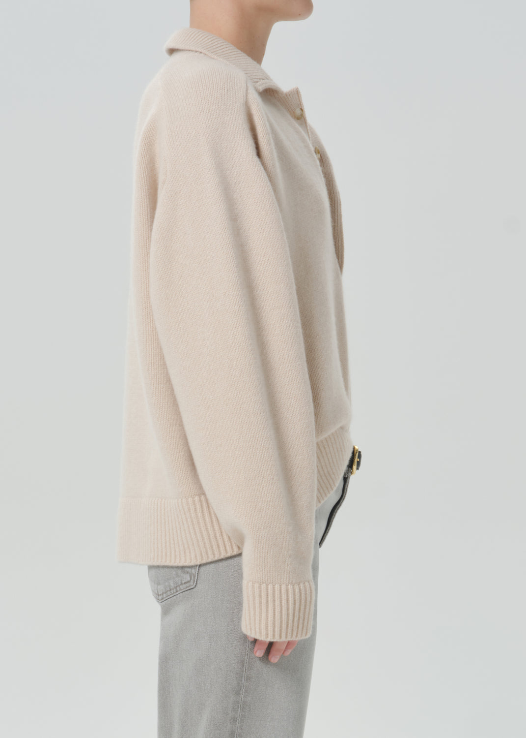 A side-profile view of a model wearing a light beige knit cardigan with a collar and ribbed cuffs, paired with light grey jeans, against a solid grey background.