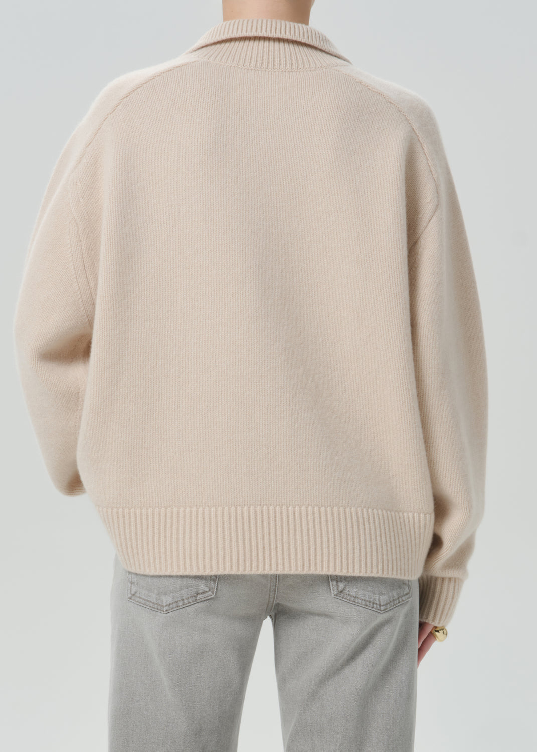 A person shown from the back wearing a light beige knit sweater with ribbed collar, cuffs, and hem, paired with light grey jeans, against a grey background.