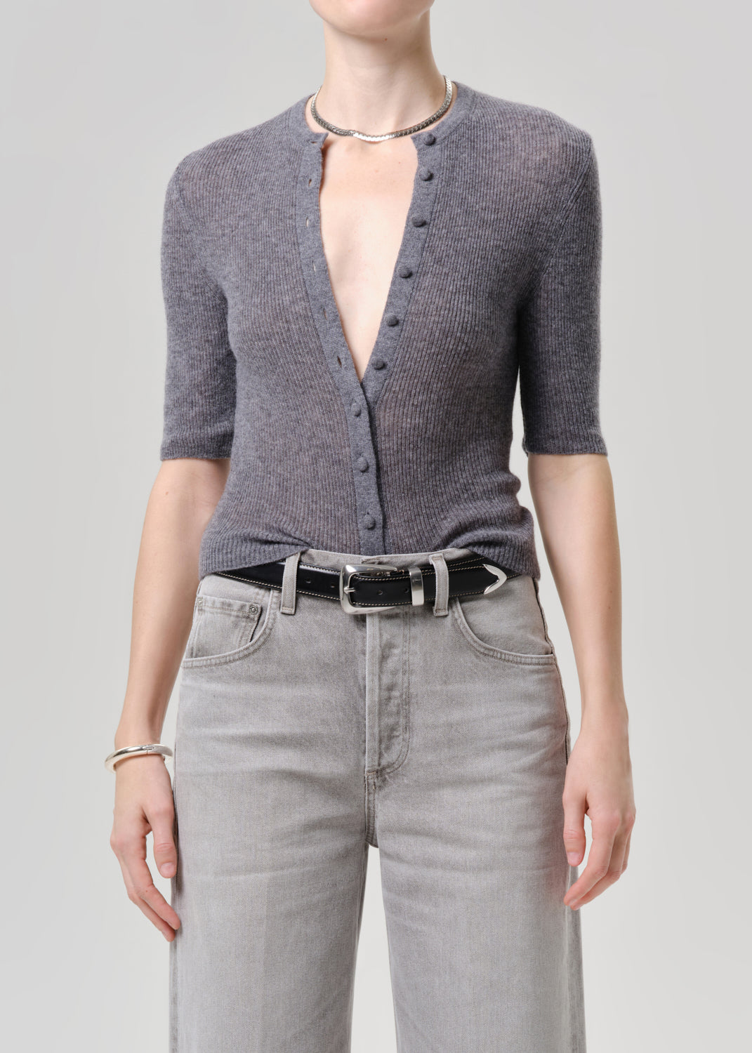A model wearing a grey short-sleeve button-front cardigan, light grey jeans, a black belt, and a silver necklace against a grey background.