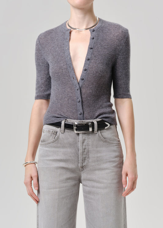 A model wearing a grey short-sleeve button-front cardigan, light grey jeans, a black belt, and a silver necklace against a grey background.