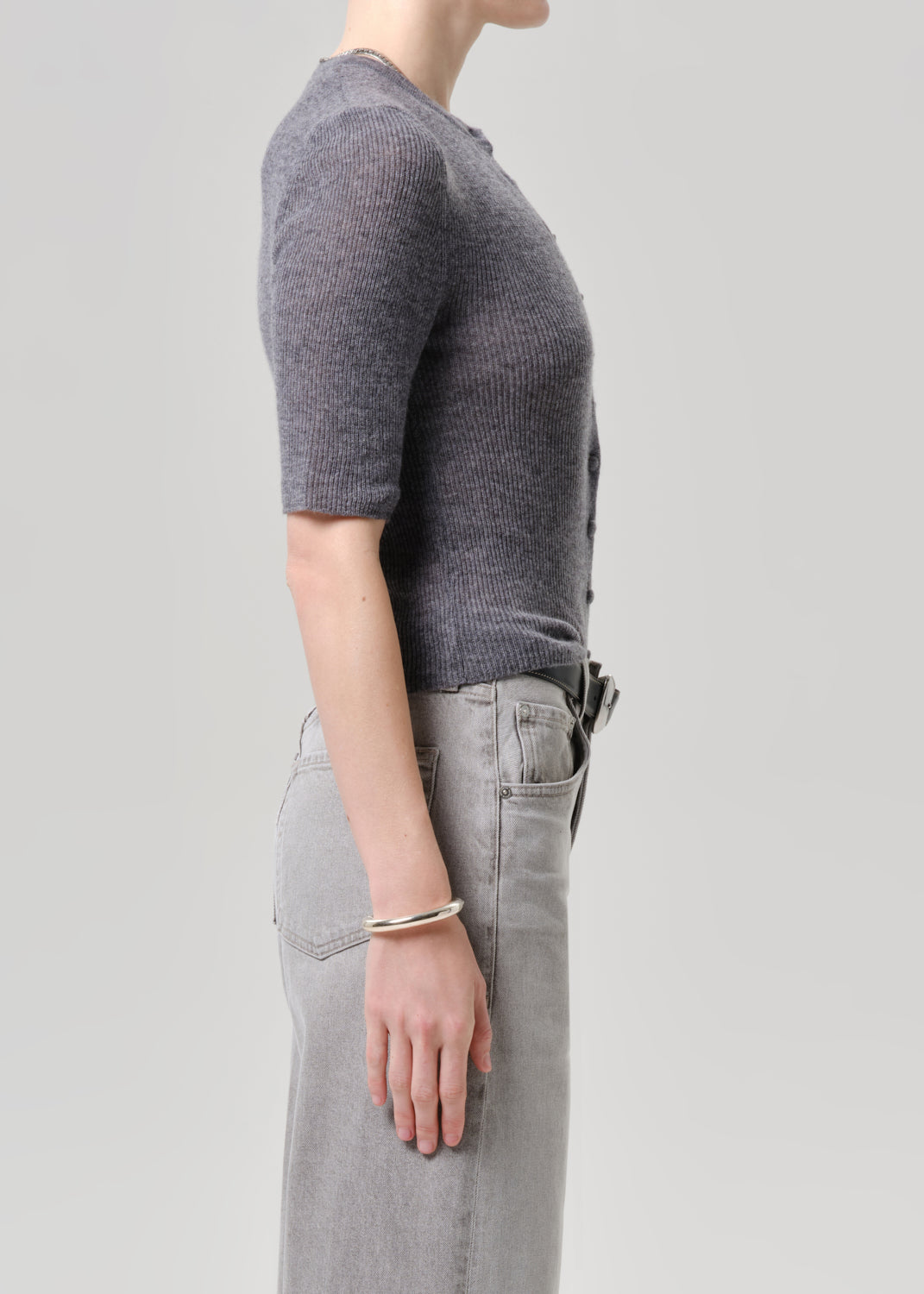 Side profile of a model wearing a short-sleeve ribbed knit top in charcoal grey and light grey jeans, accessorized with a thin silver bracelet, against a solid light grey background.