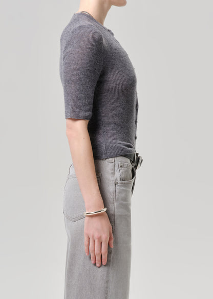 Side profile of a model wearing a short-sleeve ribbed knit top in charcoal grey and light grey jeans, accessorized with a thin silver bracelet, against a solid light grey background.