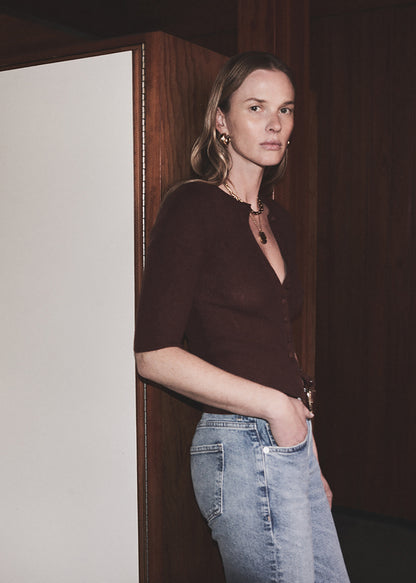 Woman wearing a dark brown top and light blue jeans standing against a wooden wall.