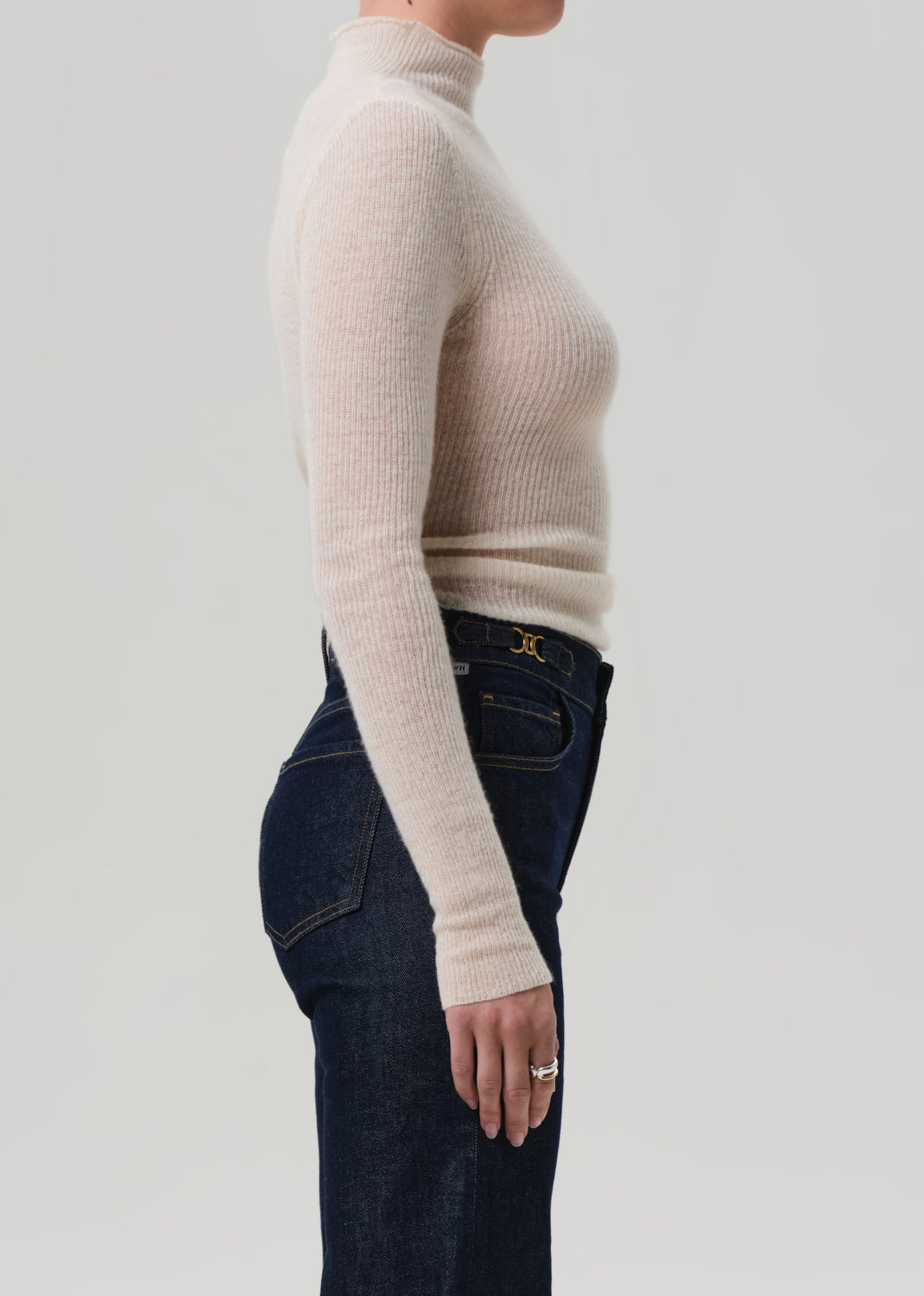Person wearing a beige turtleneck sweater and dark jeans on a light gray background