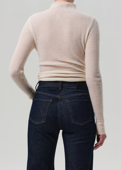 Person wearing a beige ribbed sweater and dark blue jeans on a light gray background