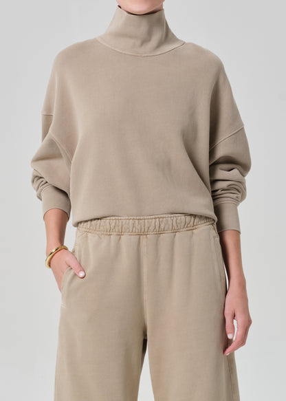 Person wearing a beige turtleneck sweater and pants on a light gray background