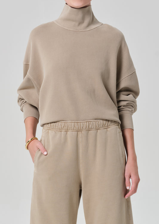 Person wearing a beige turtleneck sweater and pants on a light gray background