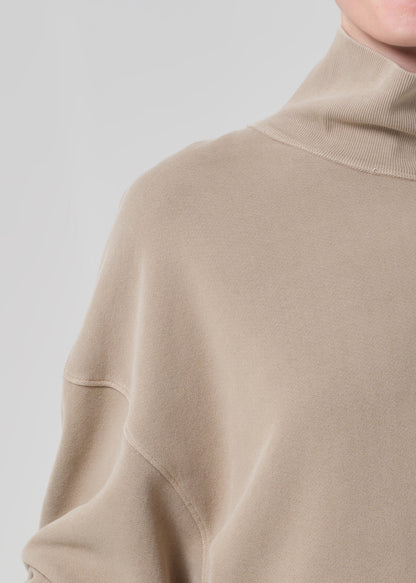 Person wearing a beige turtleneck sweater and pants on a light gray background