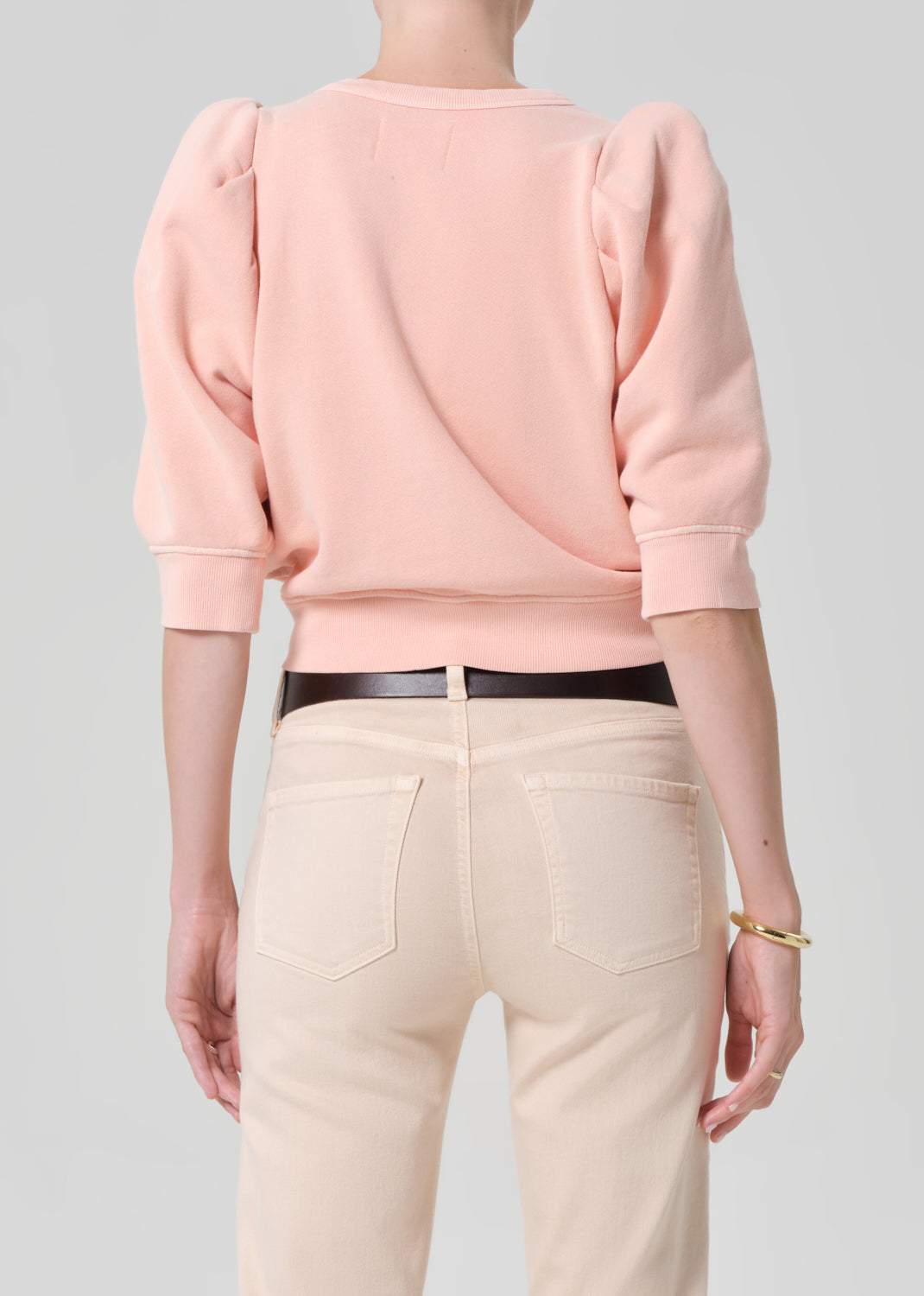 Person wearing a pink sweater and light pink pants with a belt on a gray background