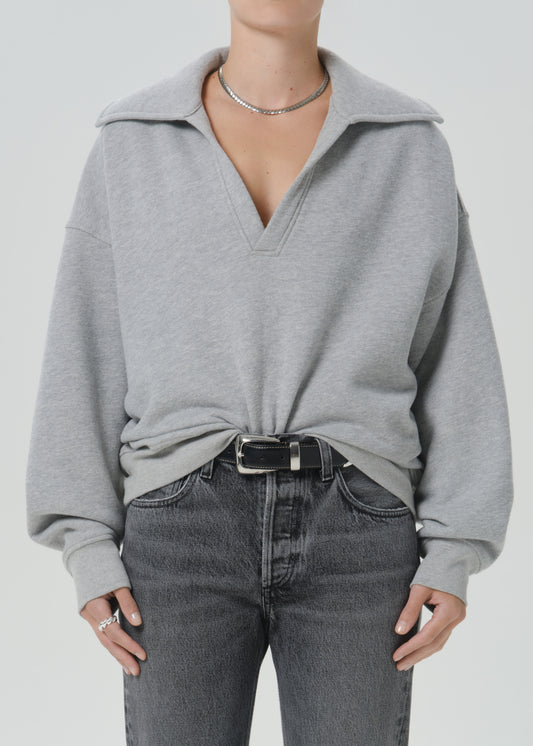 A person wearing an oversized light grey half-zip pullover sweater with dark grey jeans and a black belt, shown from the neck to the upper thighs against a grey background.
