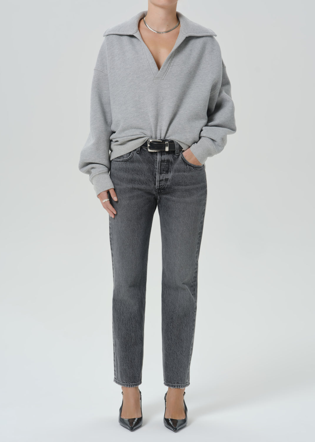 A model wearing a light grey oversized knit sweater with a collar, tucked into dark grey straight-leg jeans with a black belt, paired with black pointed-toe heels, against a grey background.