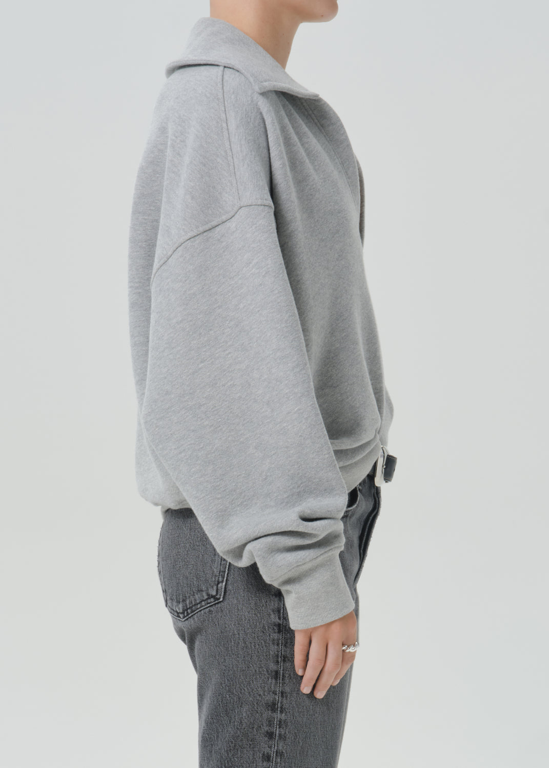 A side profile view of a person wearing a light grey oversized hoodie and dark grey jeans against a grey background.