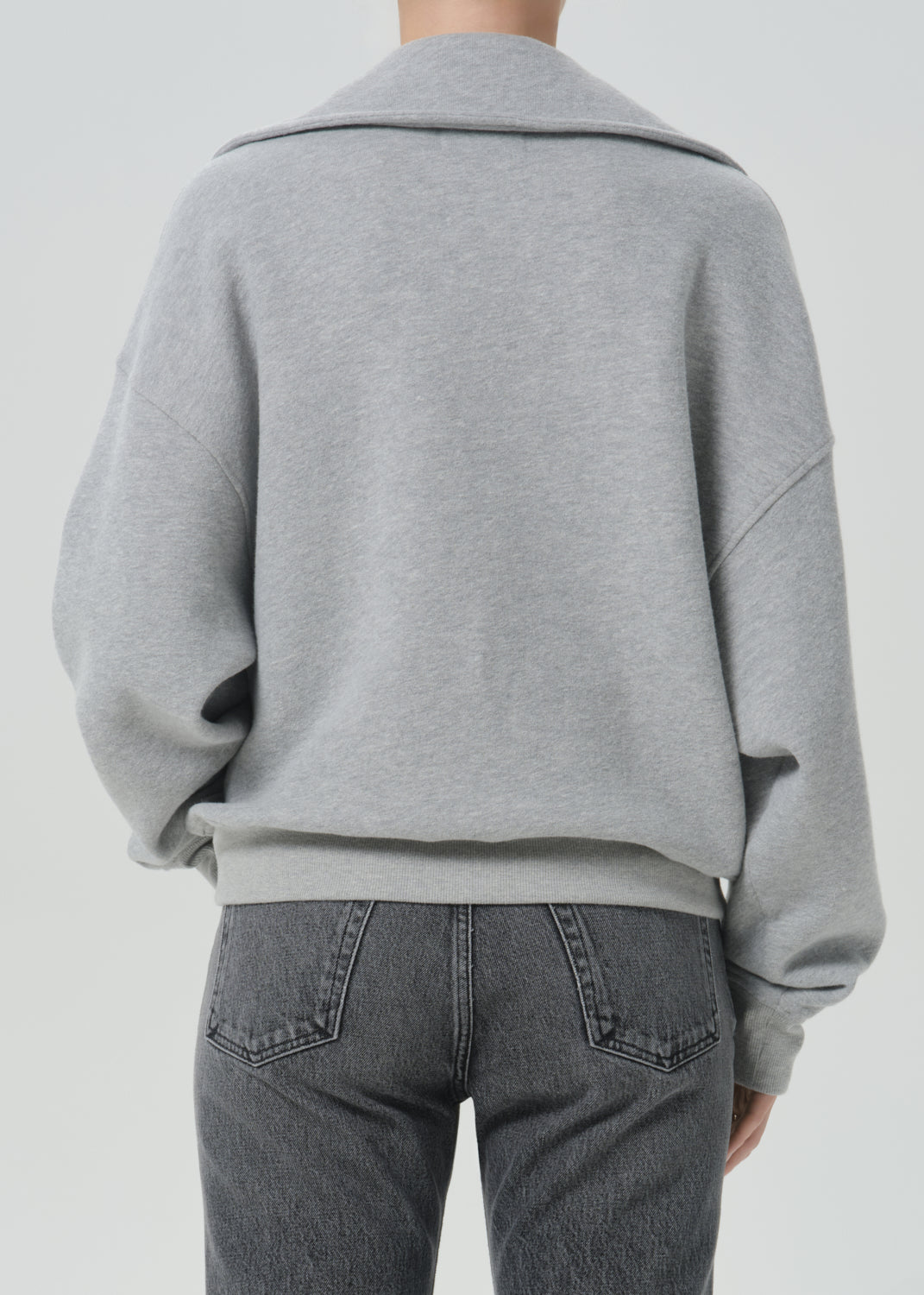 A person shown from the back wearing a light grey oversized knit top or sweatshirt with a wide collar and dark grey jeans, against a grey background.