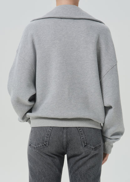 A person shown from the back wearing a light grey oversized knit top or sweatshirt with a wide collar and dark grey jeans, against a grey background.