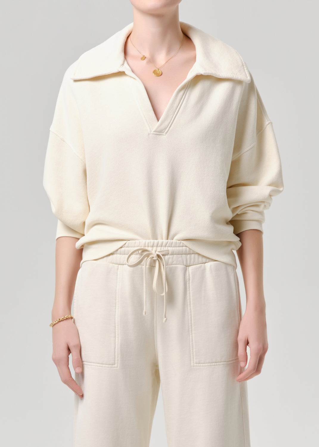A model wearing a cream-colored knit pullover top with a wide collar and matching cream drawstring sweatpants, shown against a grey background.