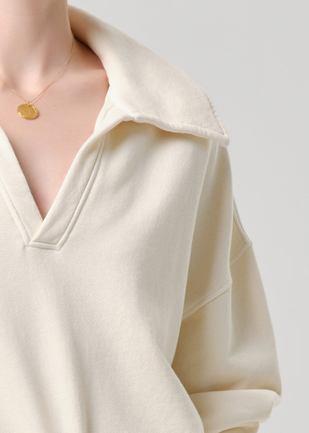 Close-up of a person wearing an oversized cream-colored knit top with a wide collar and a delicate gold pendant necklace, set against a grey background.