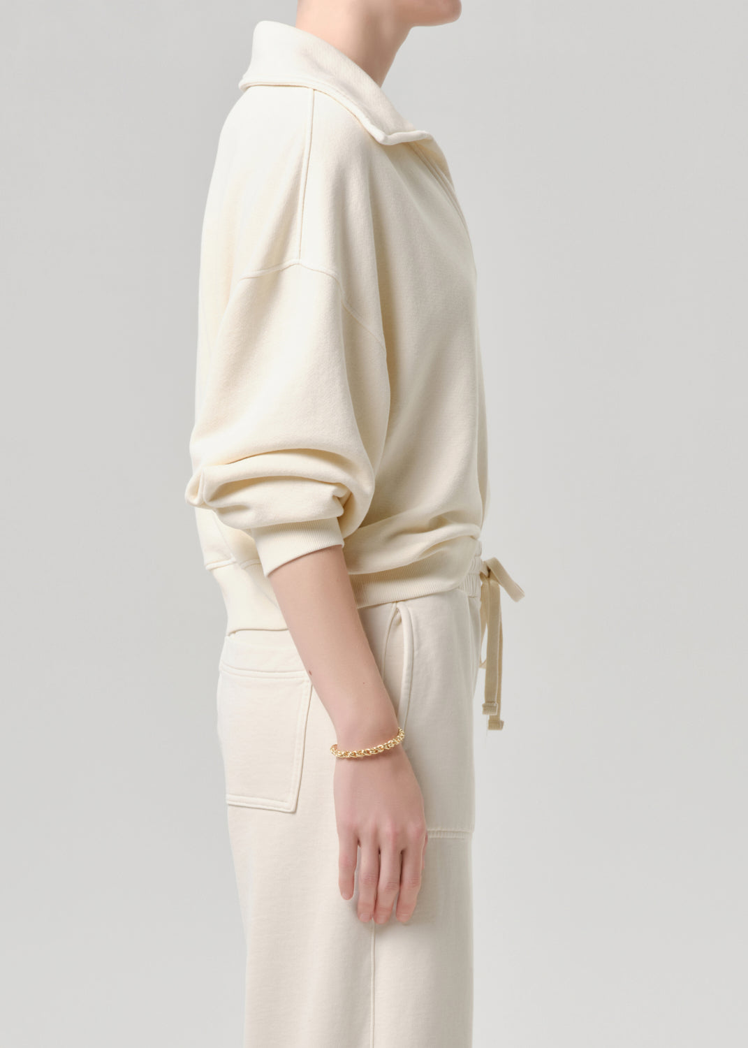 A side-profile view of a model wearing a cream-colored long-sleeve collared sweatshirt and matching cream drawstring pants against a grey background.