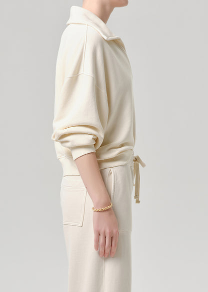 A side-profile view of a model wearing a cream-colored long-sleeve collared sweatshirt and matching cream drawstring pants against a grey background.