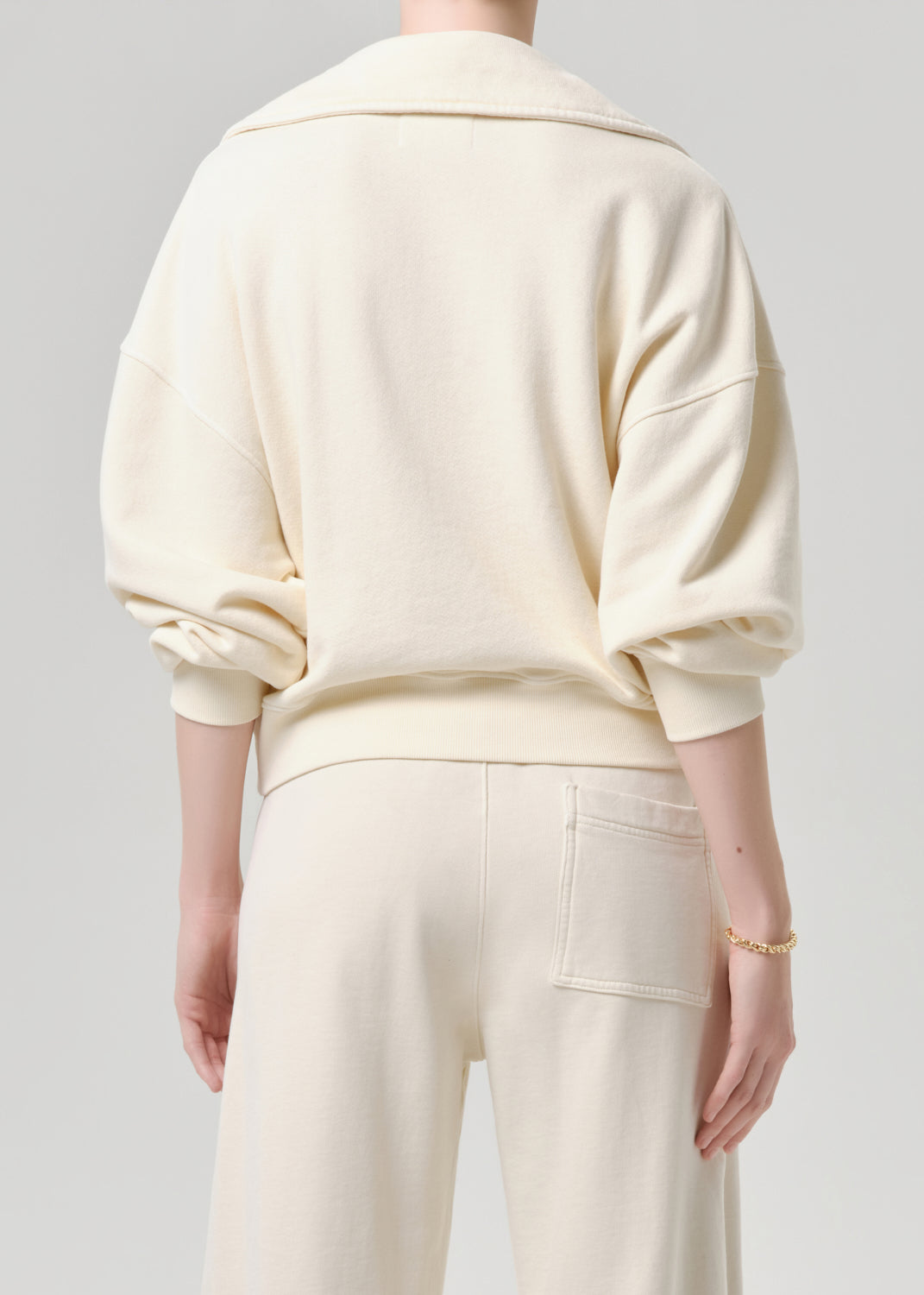 A person shown from the back wearing a cream-colored oversized sweatshirt and matching cream pants against a grey background.