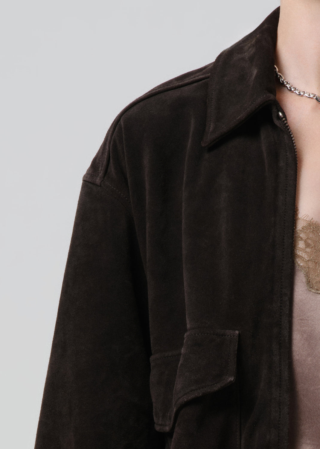 Close-up of a person wearing a dark brown suede jacket over a beige top, with a silver chain necklace visible against a grey background.