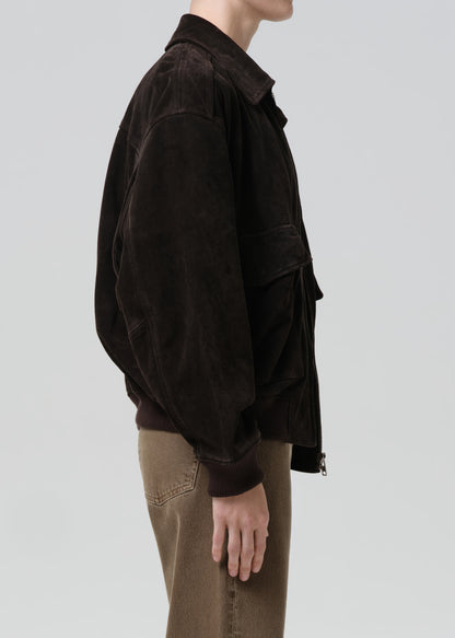 A side-profile view of a person wearing a dark brown corduroy jacket and brown pants against a grey background.