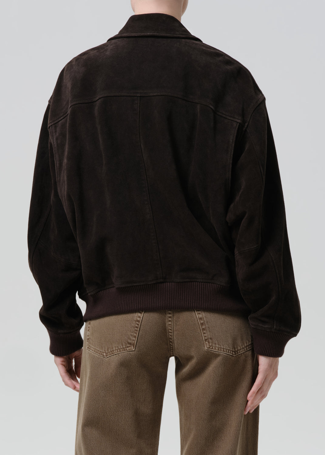 A person shown from the back wearing a dark brown suede jacket and brown pants against a grey background.