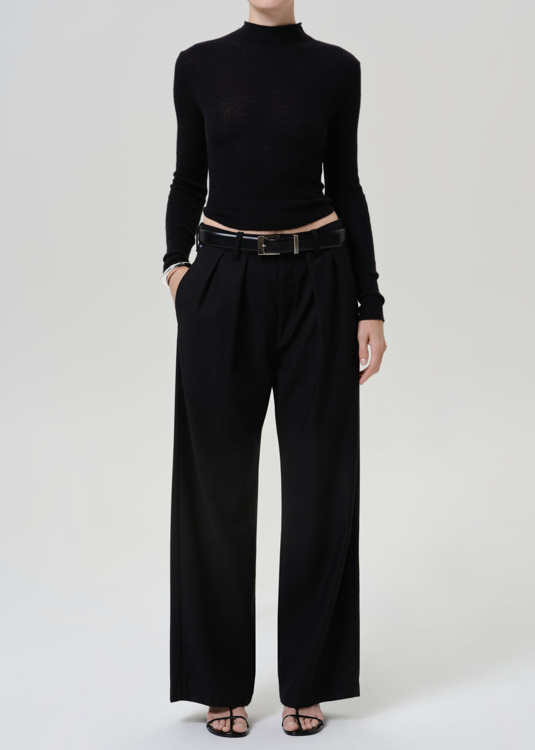A model wearing a black long-sleeve mock neck top and black wide-leg trousers, styled with a black leather belt and black flat sandals, against a light gray studio background.