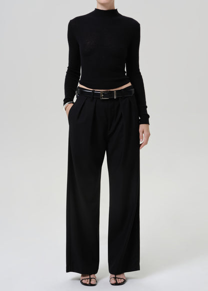 A model wearing a black long-sleeve mock neck top and black wide-leg trousers, styled with a black leather belt and black flat sandals, against a light gray studio background.