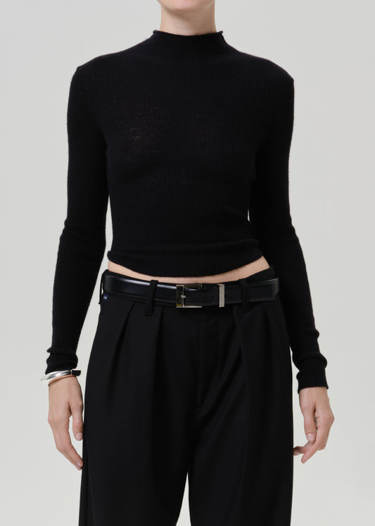 A model wearing a black ribbed mock-neck long-sleeve crop top paired with black trousers and a black belt, shown from the neck down against a light gray studio background.