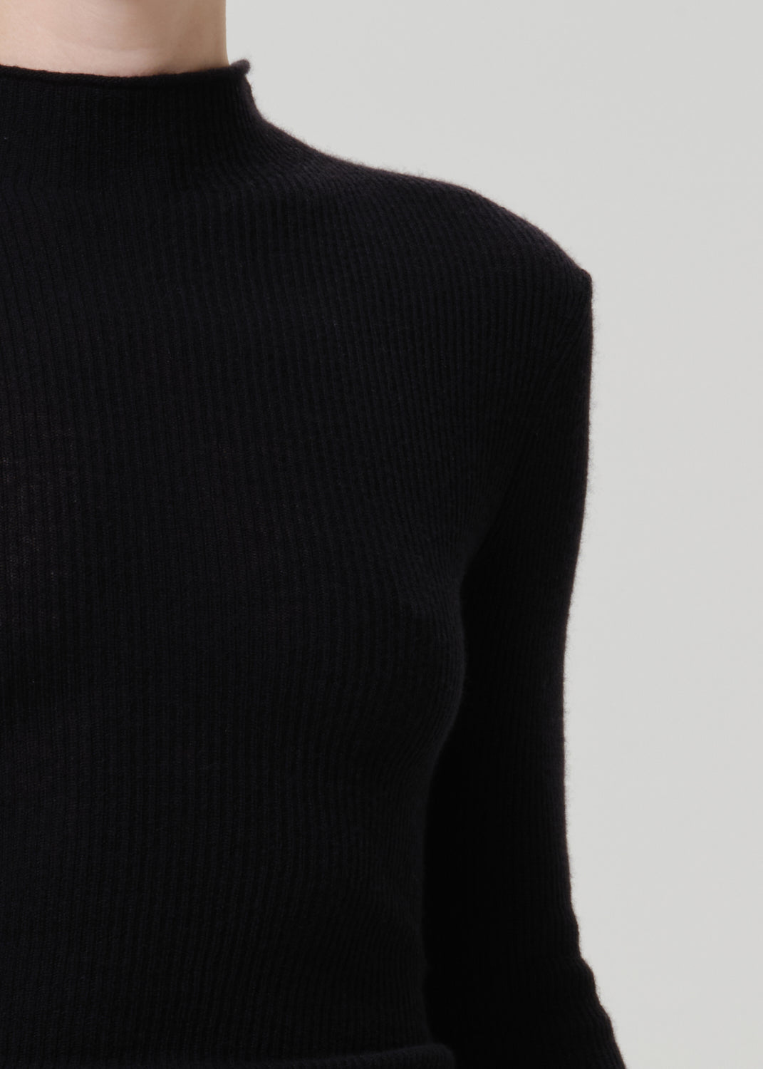 Close-up of a person wearing a black ribbed knit mock-neck sweater against a light gray background.