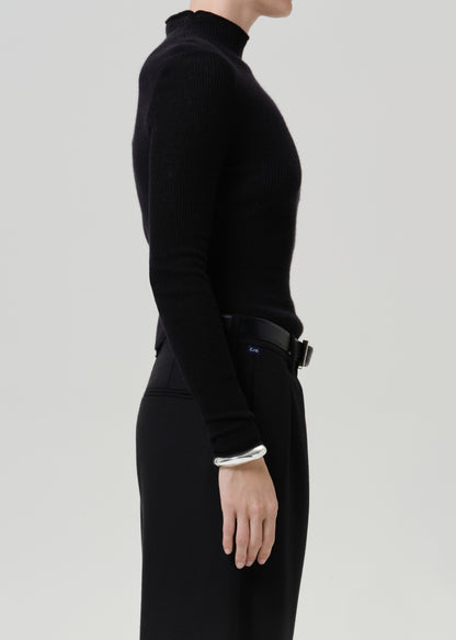 A side-profile view of a model wearing a black ribbed mock-neck long-sleeve top and black trousers, accessorized with a black belt and a silver cuff bracelet, against a light gray studio background.