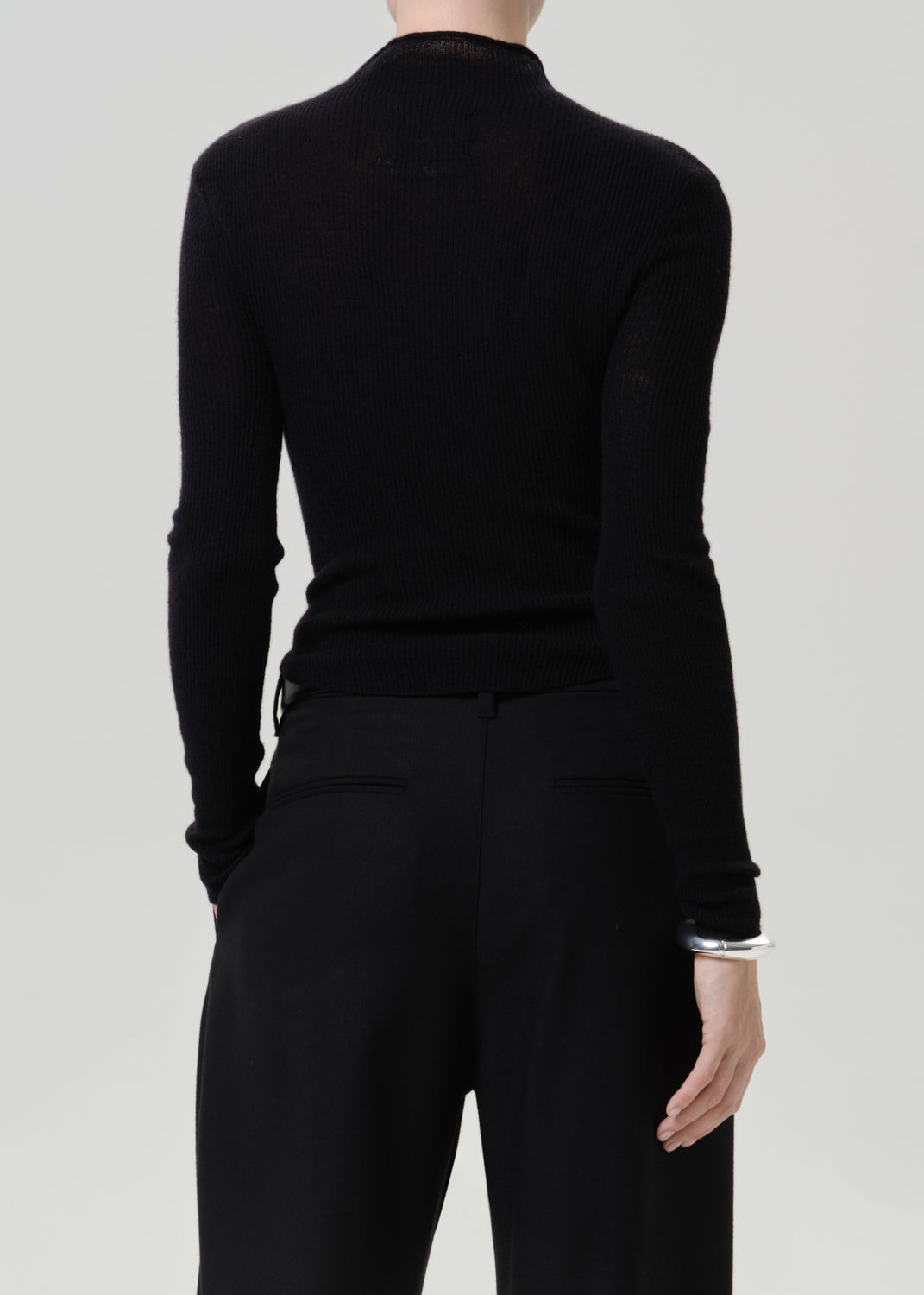 A model shown from the back wearing a black ribbed mock-neck long-sleeve top and black trousers, set against a solid light gray background.