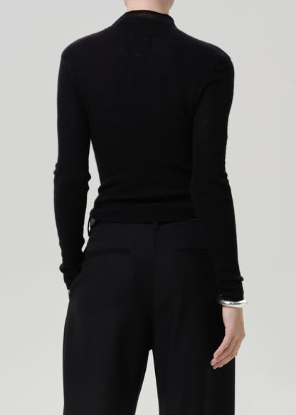 A model shown from the back wearing a black ribbed mock-neck long-sleeve top and black trousers, set against a solid light gray background.