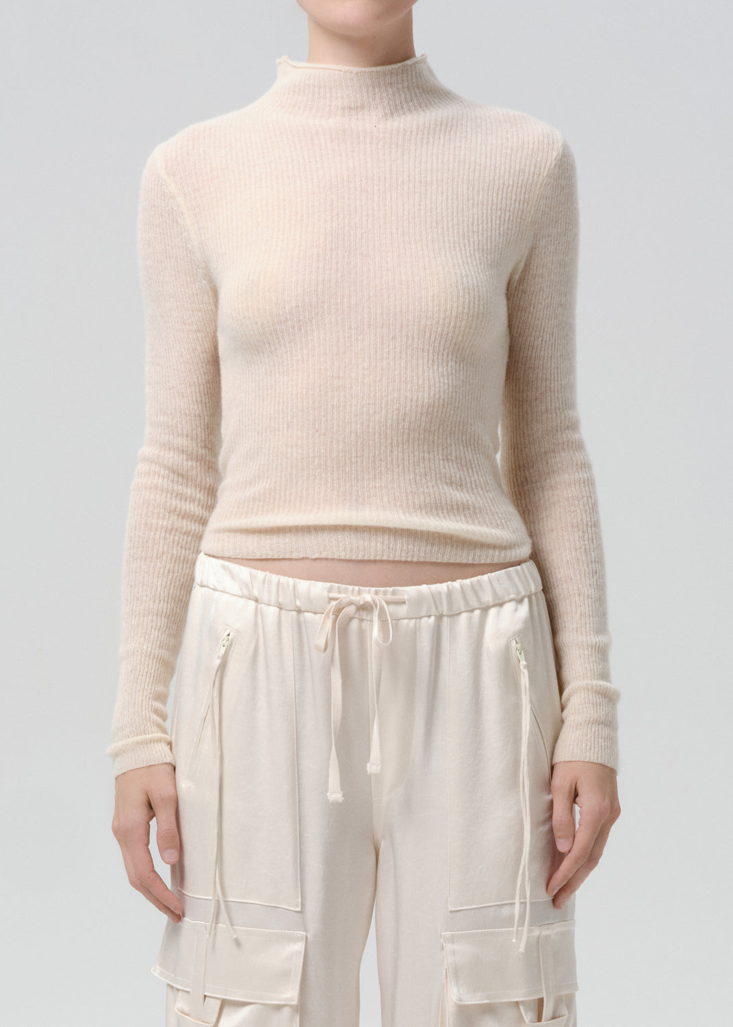 Person wearing a beige ribbed sweater and cream pants on a grey background