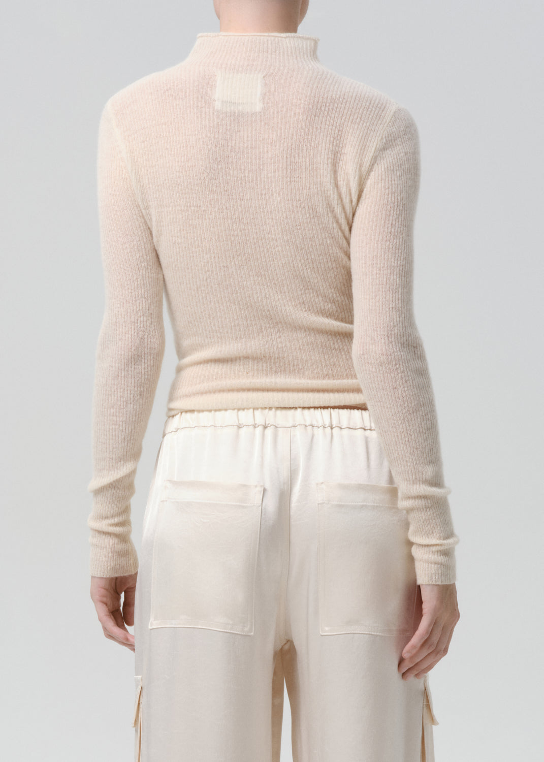 Person wearing a beige long-sleeve top and white pants on a grey background