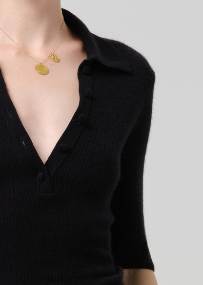 Close-up of a person wearing a black ribbed knit polo-style top with a collar and partial button placket, paired with a delicate gold necklace against a grey background.