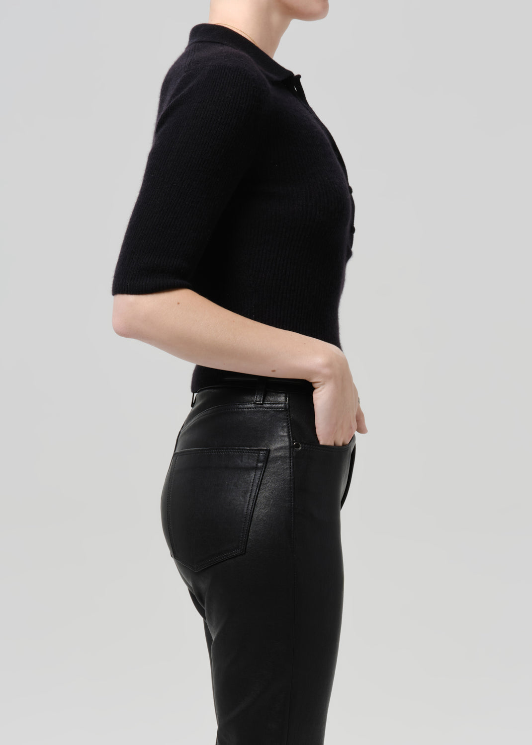 Side profile of a model wearing a black short-sleeve polo shirt and black faux-leather pants against a light grey studio background.