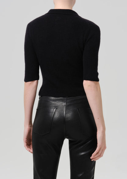 Rear view of a model wearing a black 3/4-sleeve knit polo top and black leather pants against a light grey studio background.
