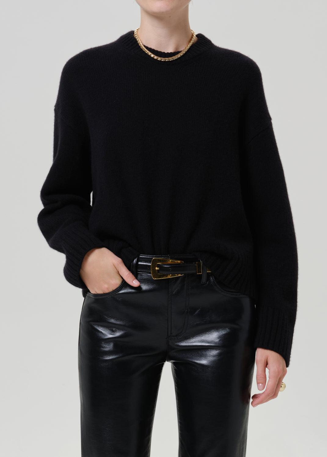 A person wearing a black knit crewneck sweater, black leather pants, and a black belt with a gold buckle, photographed against a light gray studio background.
