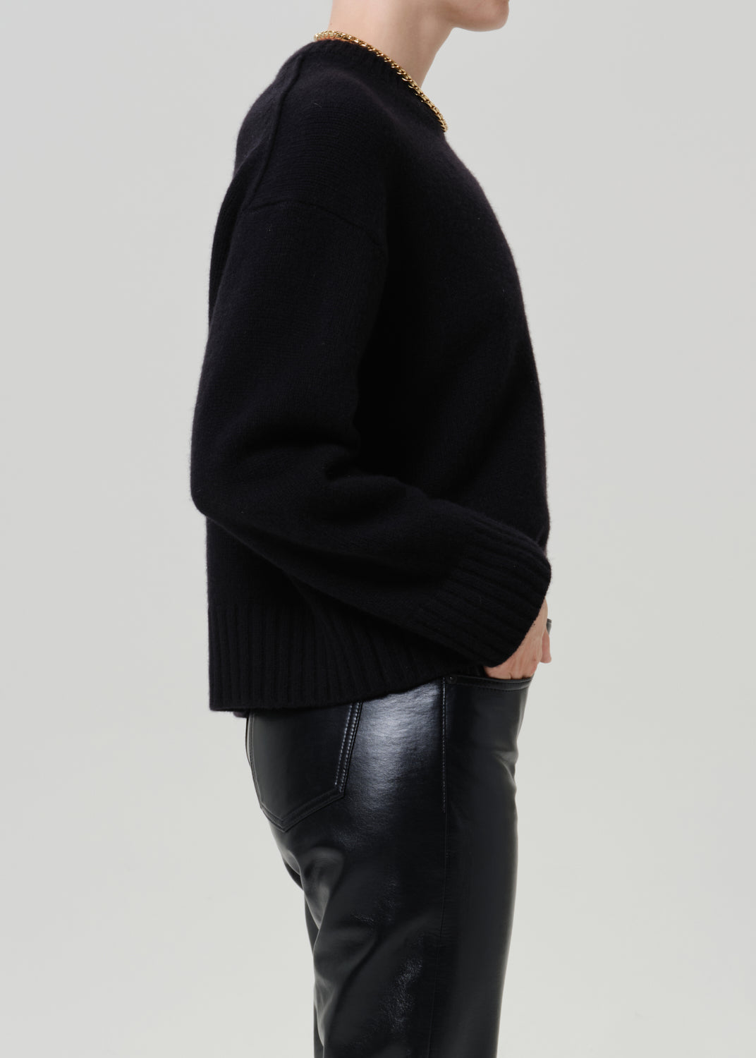A side-profile view of a person wearing a black knit sweater and black leather pants against a light gray studio background.