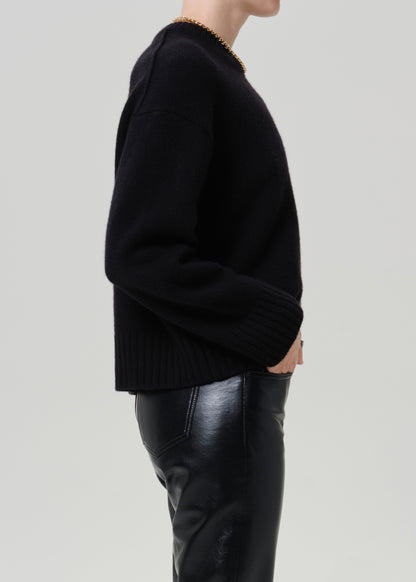 A side-profile view of a person wearing a black knit sweater and black leather pants against a light gray studio background.