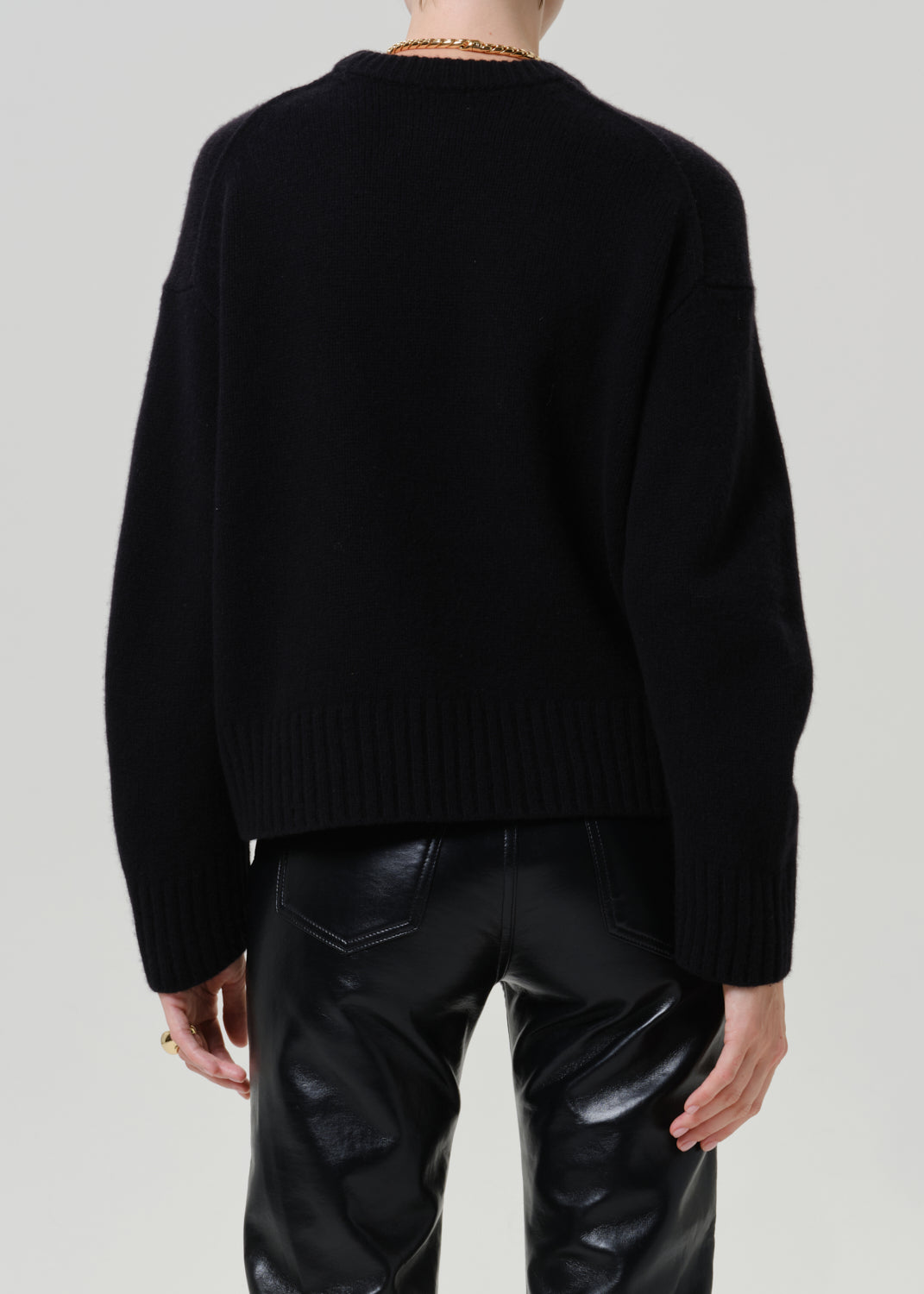A person shown from the back wearing a black knit sweater and shiny black pants against a light gray studio background.