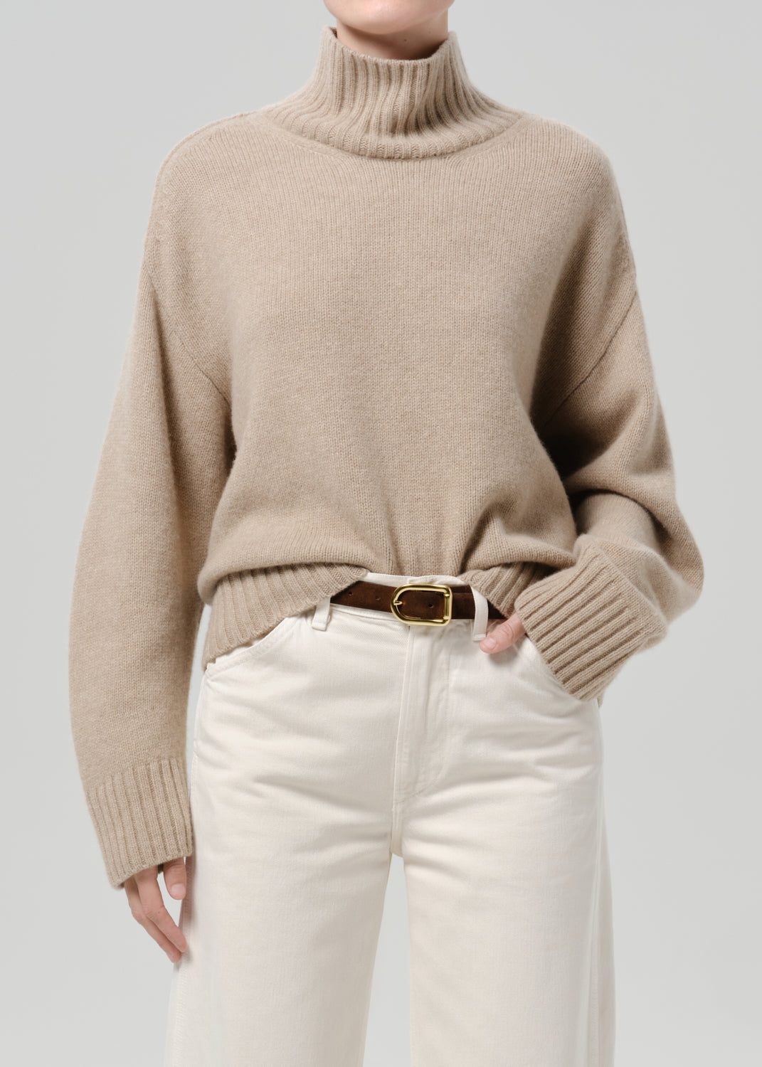 Person wearing a beige turtleneck sweater with white jeans on a grey background