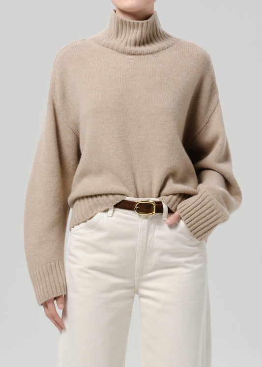 Person wearing a beige turtleneck sweater with white jeans on a grey background