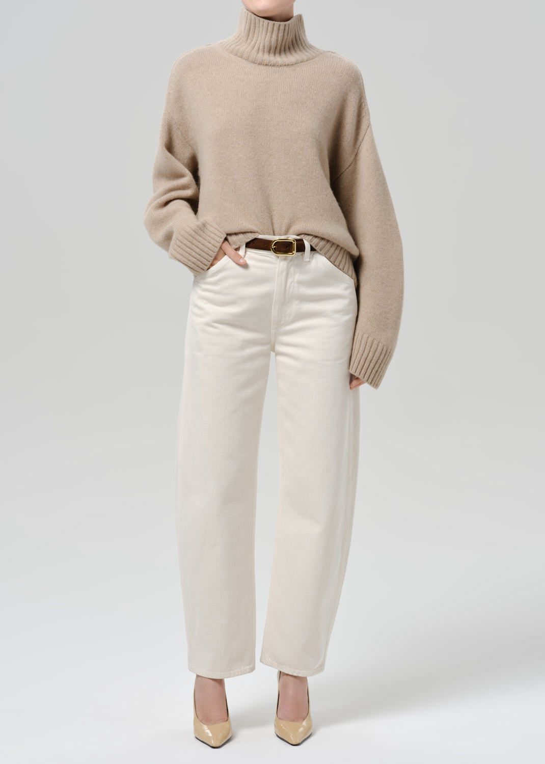 Person wearing a beige turtleneck sweater with white straight-leg jeans and beige heels on a grey background