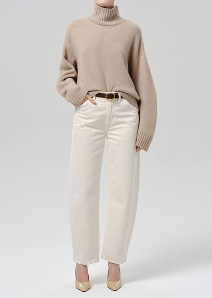 Person wearing a beige turtleneck sweater with white straight-leg jeans and beige heels on a grey background