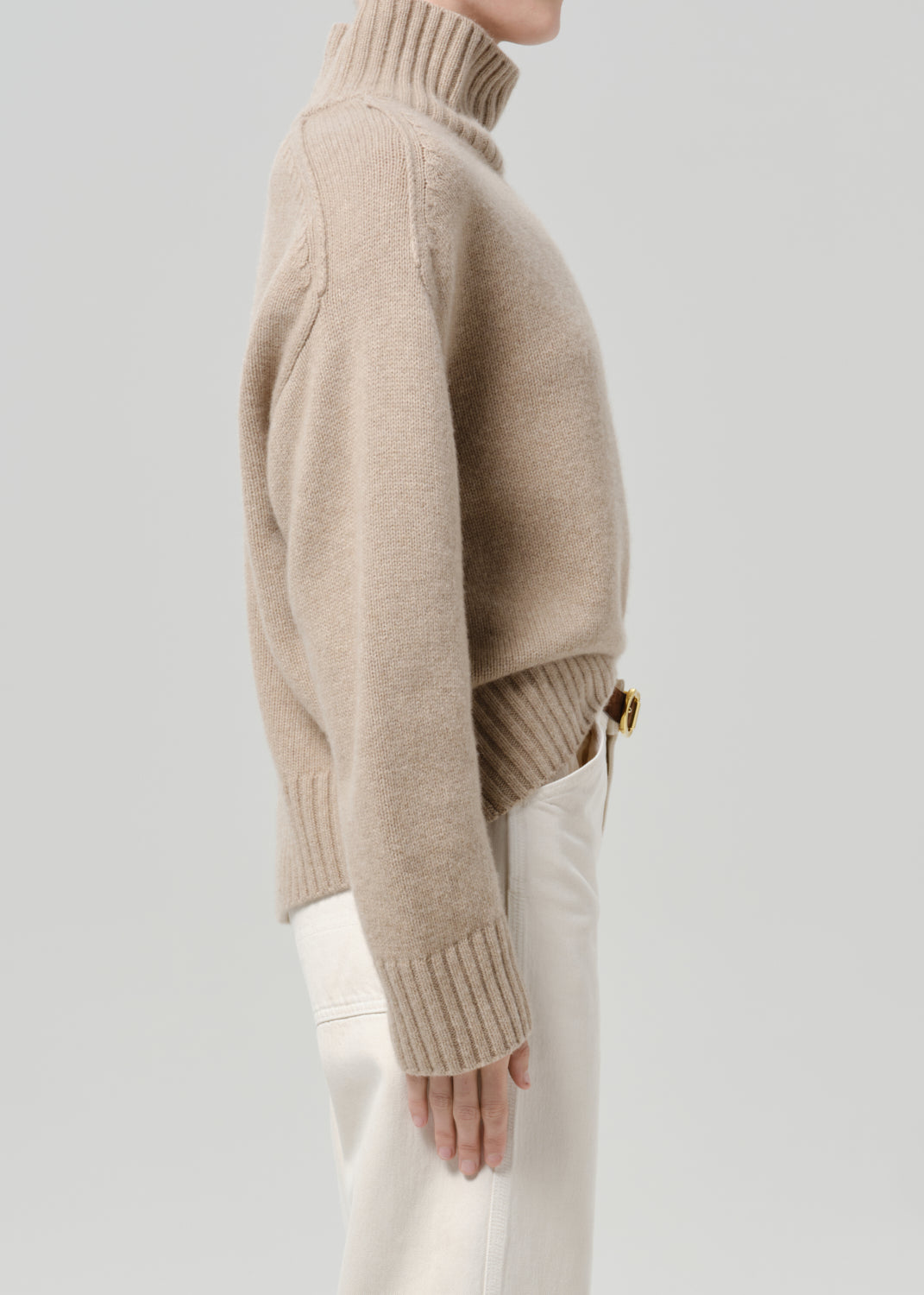 Person wearing a beige turtleneck sweater with white jeans viewed from the side on a grey background