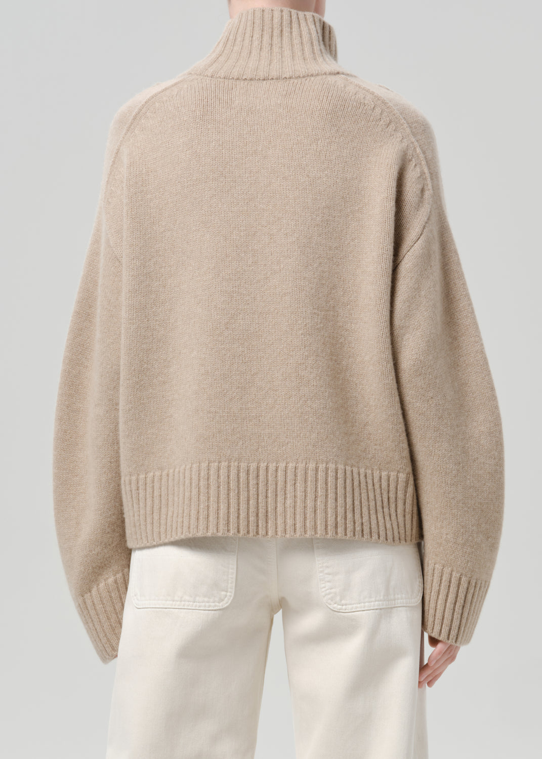 Person wearing a beige turtleneck sweater with white jeans viewed from the back on a grey background