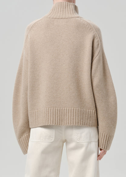 Person wearing a beige turtleneck sweater with white jeans viewed from the back on a grey background