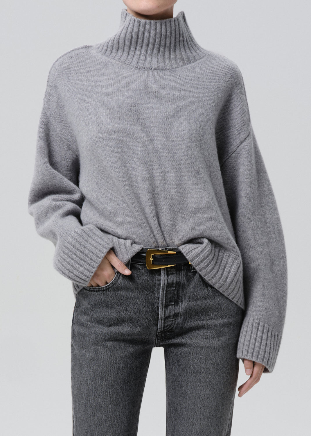 Person wearing a light grey oversized turtleneck sweater with grey straight leg jeans on a grey background